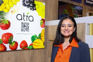 A smiling woman with dark shoulder-length hair is standing in front of a wall on which fruit can be seen.