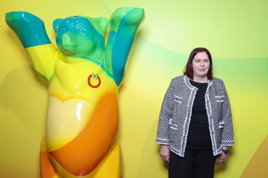 Frau steht neben einer bunten Bärenfigur vor gelbem Hintergrund.