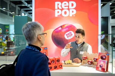 On a trade fair stand Alexander Höller talks about the apple he is holding to two trade visitors with their backs to the camera.