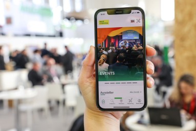 A photograph showing a hand holding a smartphone, in the background people in the exhibition halls.