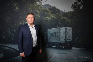 Photo of a man in front of a screen, a lorry in the background.