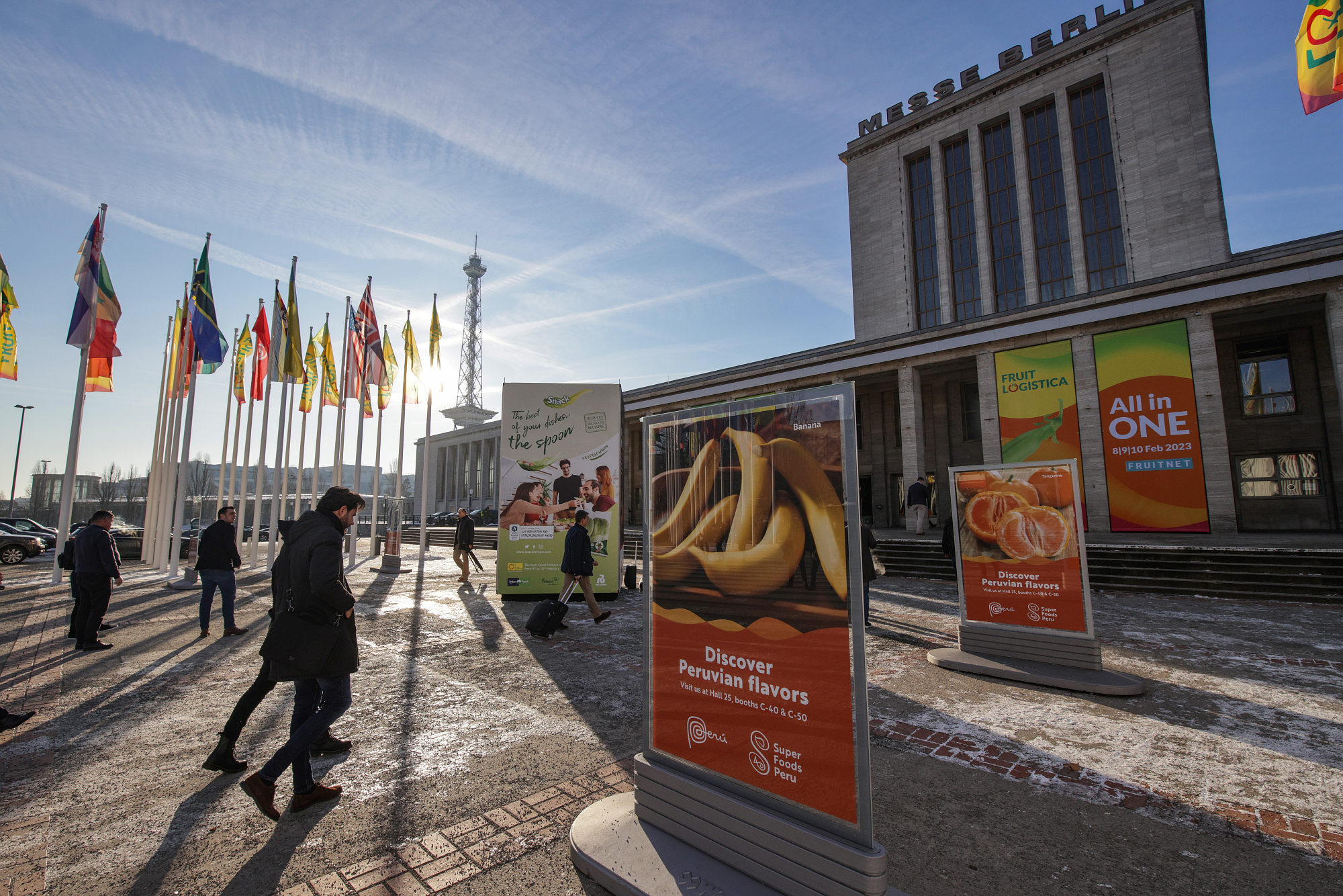 The photo shows a long shot of the Messe Berlin North Entrance with country flags and FRUIT LOGISTICA 2023 displays. People are walking by.
