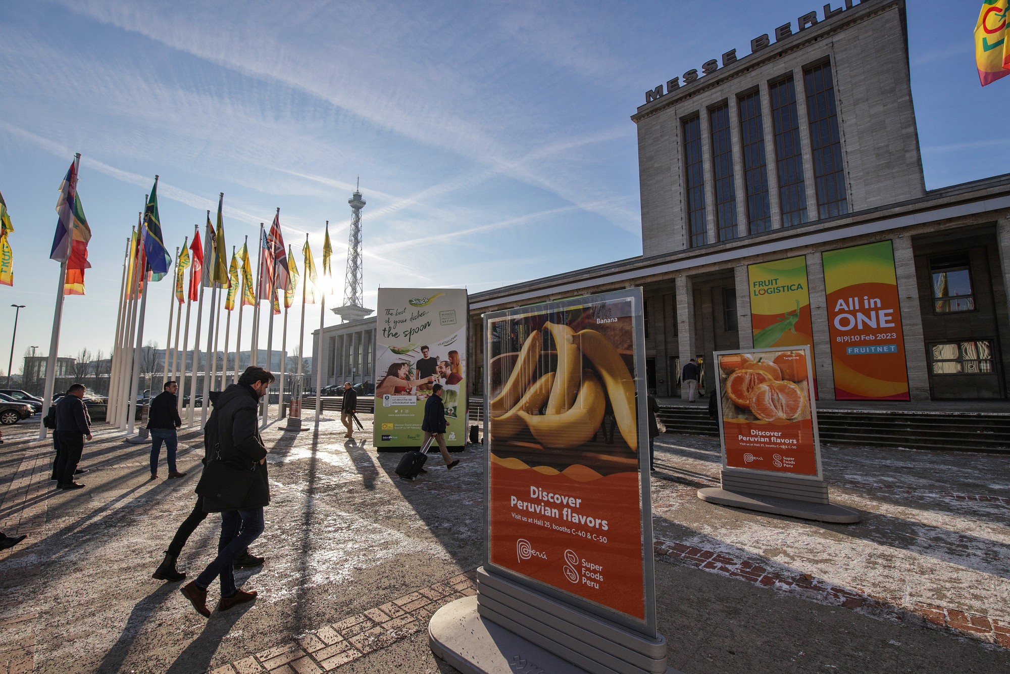 Das Foto zeigt eine Totale des Messe Berlin Nordeingangs mit Länderflaggen und Aufstellern der FRUIT LOGISTICA 2023. Menschen laufen vorbei.