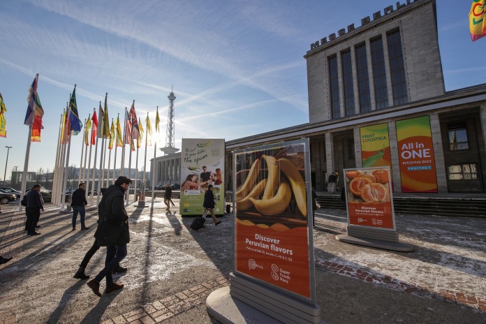 The photo shows a long shot of the Messe Berlin North Entrance with country flags and FRUIT LOGISTICA 2023 displays. People are walking by.