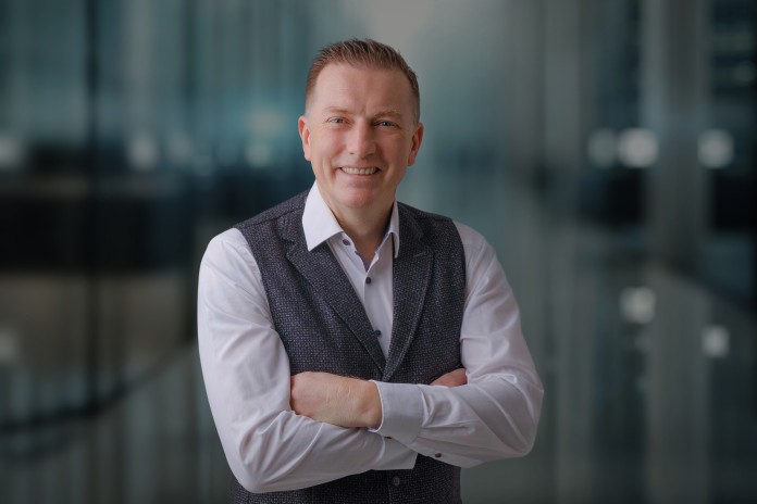 A man in a modern office building. He is wearing a white shirt and a dark waistcoat. His arms are crossed in front of his chest as he smiles into the camera. 